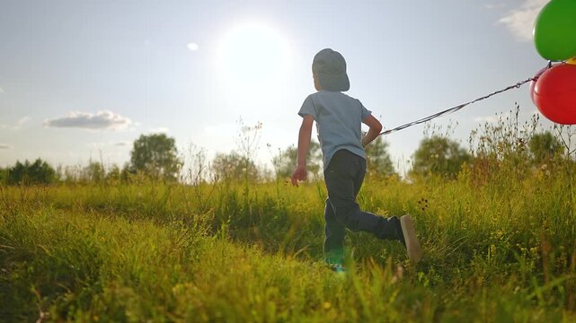 Boy pulls colorful balloons while running through meadow field. Child with balloon bunch runs in wildflower meadow. Kid pulling balloons outdoors. Boy runs with colorful balloons in sunny field.