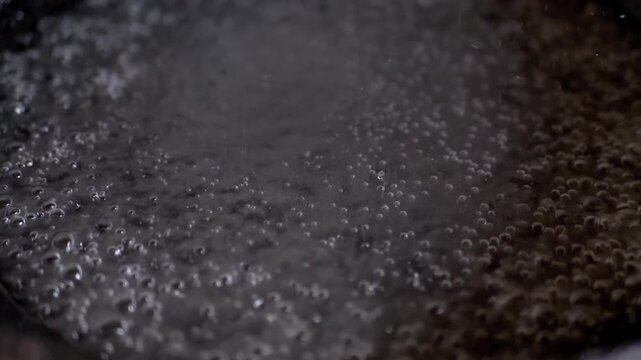 An intense close-up shows water rapidly boiling in a metallic pot, with steam rising and countless small bubbles forming and bursting. Captures the energy of heating liquid.