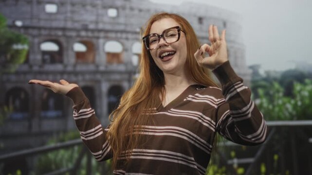 Redhead woman in striped sweater and glasses holds ok sign with right hand and open palm toward an ancient building colosseum in rome; joy travel.
