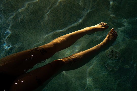 Close-up of a person's legs and feet partially submerged in blue pool water with atmospheric low light and reflections. Concept of relaxation, spa, and wellness vacation.