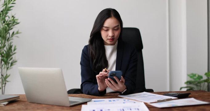 Asian businesswoman having a phone conversation while analyzing financial reports and using a laptop at her rustic wooden desk, demonstrating productivity and multitasking in a professional setting