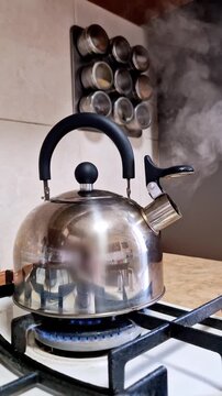A stainless steel kettle boils on a gas stove with a bright blue flame visible underneath the pot. Steam steadily rises from the spout as the water heats up in a modern kitchen setting for a hot drink