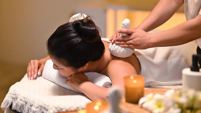 Woman receiving herbal compress massage treatment at spa, therapist applying hot compress on back in warm relaxing atmosphere.
