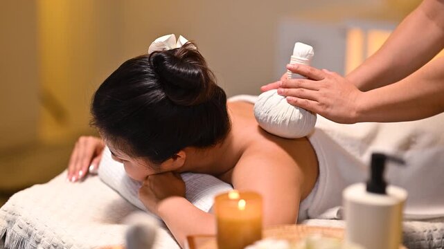 Woman receiving herbal compress massage treatment at spa, therapist applying hot compress on back in warm relaxing atmosphere.
