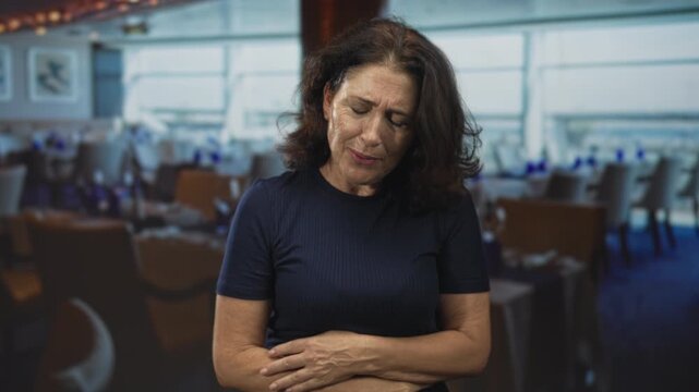 Woman holding hands over abdomen and leaning forward in building dining area with set tables, chairs and large windows visible; pain vulnerability.