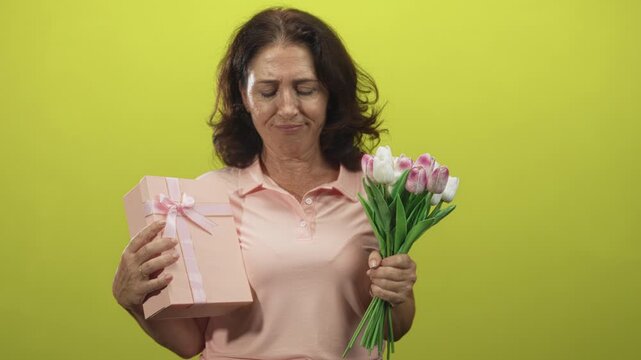 Woman holding pink gift box and bouquet with hands, eyes closed and slight grimace in studio; bittersweet memory reflection.