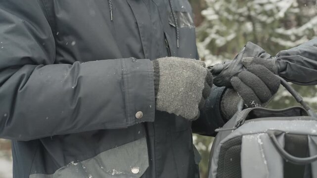 Hands packing backpack in snow, gray gloves and waterproof jacket, zipper being closed, snowflakes settling on fabric, preparing camera and laptop gear, careful organization and protective layering,