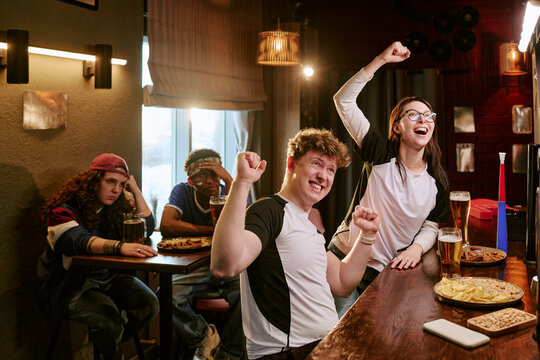 Young adult sports fans celebrating match result in bar while disappointed friends watching nearby, showing contrasting emotions. Ideal for sports marketing, fan culture, teamwork campaigns