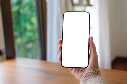 Mockup image of a woman holding mobile phone with blank white desktop screen at home