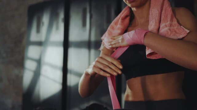 Woman wrapping hands with boxing bandages in gym with towel over shoulders, preparing for intense workout focused on strength and endurance