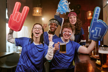 Obraz premium Portrait of diverse young adult sports fans cheering in bar, raising foam fingers and beer mugs during match viewing. Ideal for fan culture, team support, sports marketing campaigns