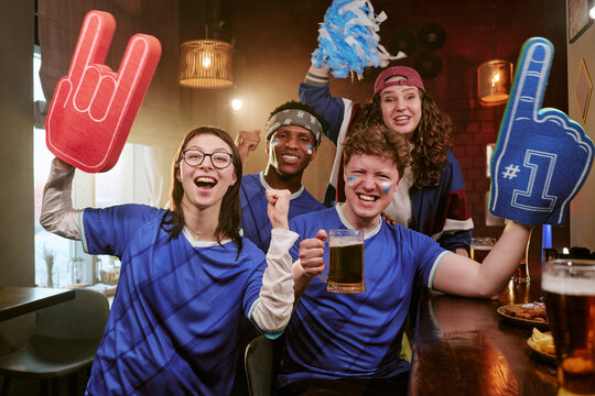 Portrait of diverse young adult sports fans cheering in bar, raising foam fingers and beer mugs during match viewing. Ideal for fan culture, team support, sports marketing campaigns