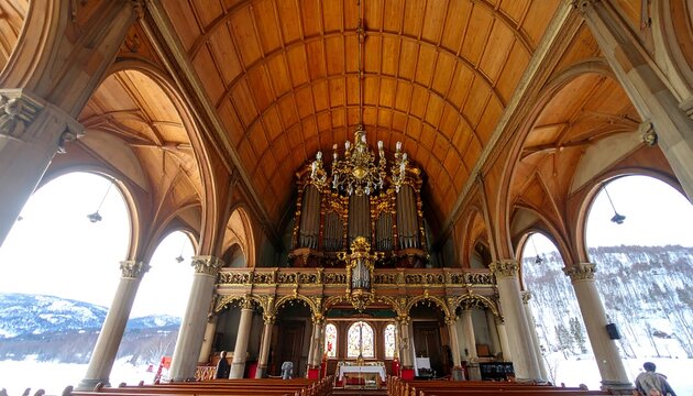 Interior view of a church with high arched ceiling and ornate pipe organ, visible light streams, and a glimpse of a snowy landscape