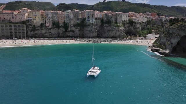 Drone orbit around catamaran in Tropea bay with cliffside town view, Italy
