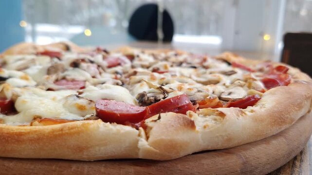 Whole pizza with sausage, mushrooms, and melted cheese rests on a wooden serving board. Shallow focus shot reveals the appetizing texture with a bright, blurred window background.
