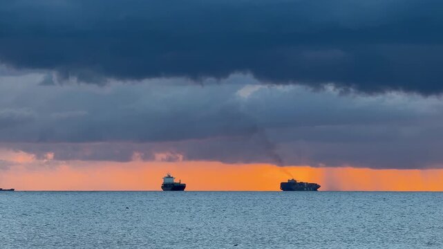 Two large ships move across the ocean as the sun sets on the horizon. Clouds hang above the water and the light reflects on the waves. This moment shows a vast ocean view.
