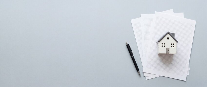 A small white house model sits on top of stacked papers next to a pen on a gray surface.