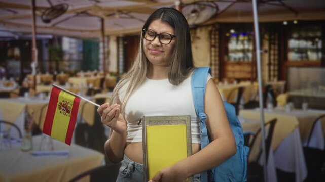 Young woman holding a spanish flag and textbooks with backpack, grimaces while standing in a street cafe; study frustration.