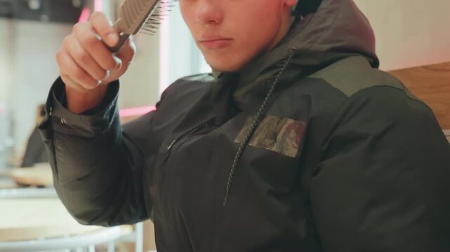 Young man combing hair in cafe, metal comb pulled from backpack, seated on wooden bench, winter jacket on, focused expression, soft neon lighting, quick onthego grooming moment, practical selfcare