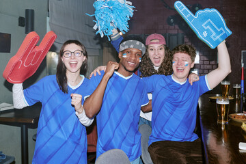 Obraz premium Portrait of multiethnic young adult sports fans cheering in bar, raising foam fingers and pom poms, celebrating team win. Ideal for fan culture, sports marketing, group loyalty campaigns