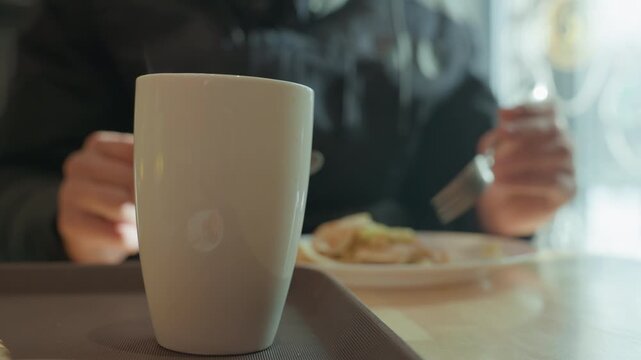 White mug foreground person eating breakfast soft sunlight through window ceramic cup in sharp focus blurred diner reaching with fork plate of eggs and toast on tray intimate cinematic closeup warm