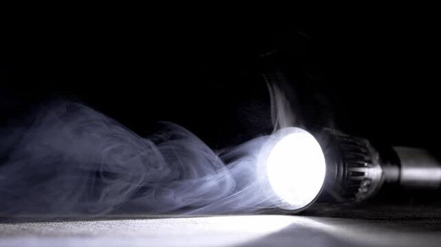 A bright LED flashlight lies on a dark surface, casting a strong beam through thick swirling white smoke. Concept of mystery, emergency search, power outages, and finding direction.