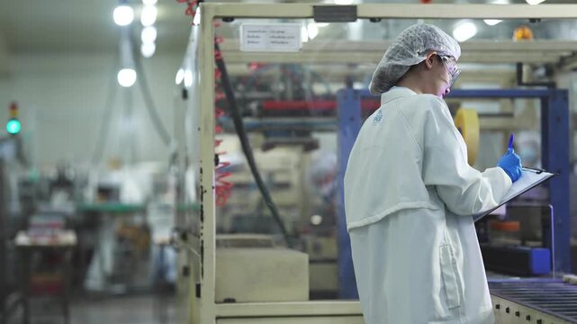 A focused female quality inspector in a sanitized lab coat and safety glasses recording data while auditing an automated production line in a modern manufacturing facility.