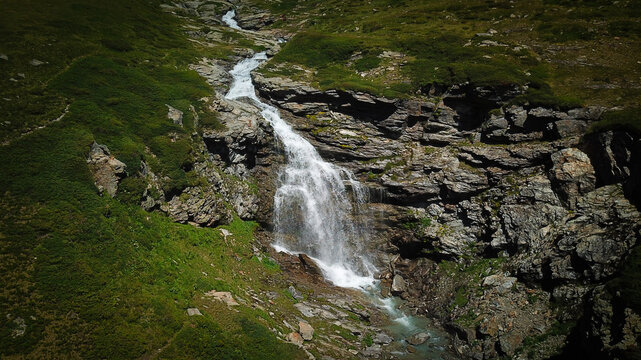 Aerial view of a cascade of water tumbling over the rugged, dark rocks, contrasting against the vibrant green slopes, Valgrisenche, Aosta Valley, Italy.