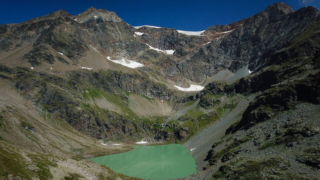 Aerial view of Lago di San Grato nestled amidst rugged, rocky terrain and snow-capped peaks under a clear blue sky, Valgrisenche, Aosta Valley, Italy.