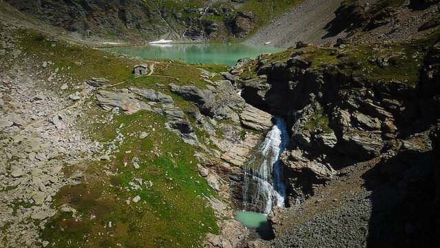 Aerial view of the cascade's waters plunging into a turquoise pool below, against the backdrop of Lago di San Grato, Valgrisenche, Aosta Valley, Italy.
