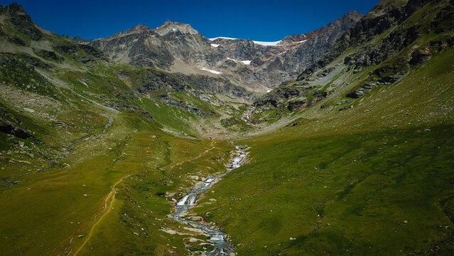 Aerial view of a meandering stream cutting through verdant meadows towards rugged, snow-capped peaks, Lago di San Grato, Valgrisenche, Aosta Valley, Italy.