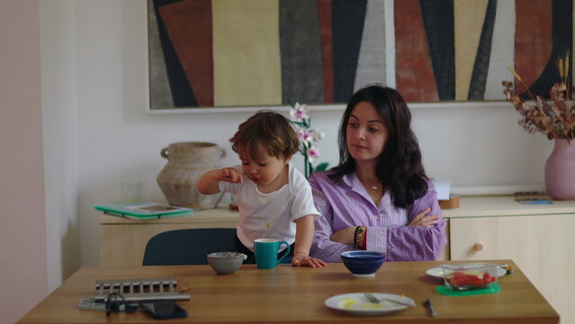 Mother watching toddler refuse breakfast while sitting at table with crossed arms, child playing with spoon and food, candid parenting moment showing patience and everyday family dynamic