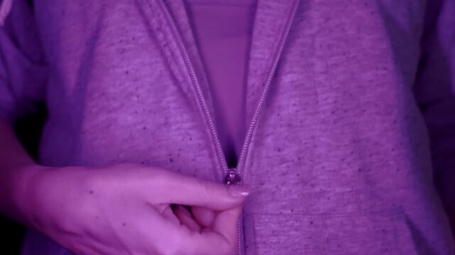 A close-up view captures a woman's hand slowly pulling down the zipper of a grey hooded sweatshirt. Concept of undressing, casual fashion, and revealing layers in dramatic purple lighting.