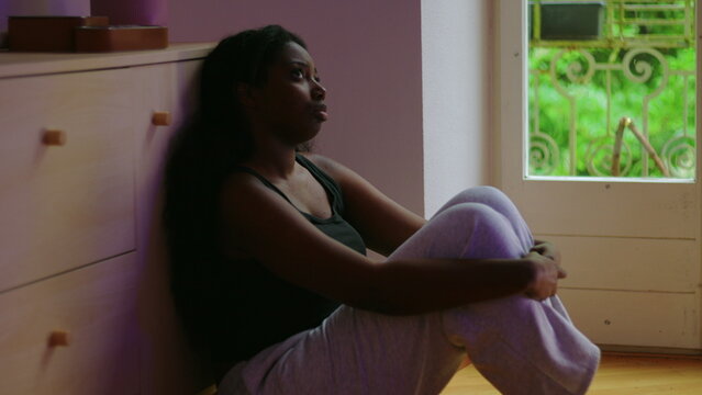 African American woman sitting on floor by window hugging knees looking ahead with distant expression showing introspection worry and aimlessness during difficult life moment