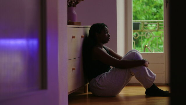 African American woman wide shot sitting on floor by window leaning against cabinet in still posture showing introspection worry and aimless reflection in quiet home space