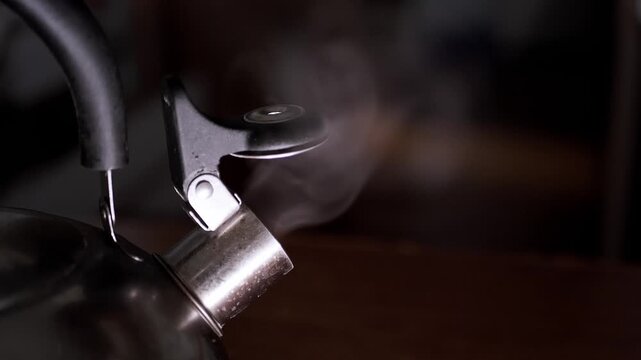 Thick hot steam billows from the open spout of a stainless steel tea kettle boiling in a dark room. Concept of cozy morning routines, making warm beverages, and relaxing home life.