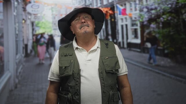 Senior man fisherman wearing wide brim hat and cargo fishing vest sticks tongue out while posing in a cobbled street with shops and flags; playful curiosity.