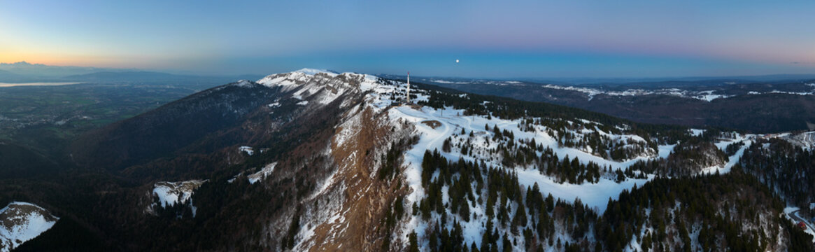 Aerial view of snow-dusted peaks and verdant forests meet under a pastel sky, crowned by a solitary tower, Petit Mont Rond, Auvergne-Rhone-Alpes, France.