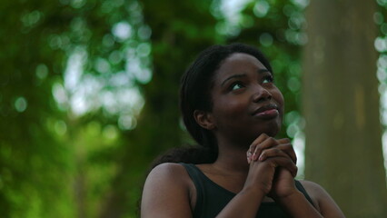 Fototapeta premium African American woman standing outdoors raising hands and looking upward before closing eyes and bringing hands together in prayer creating spiritual moment of hope and faith in natural environment