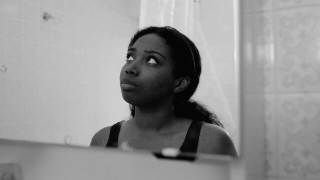 African American woman looking down at her reflection in bathroom mirror with sad and troubled expression creating emotional monochrome moment of self doubt and vulnerability in indoor setting