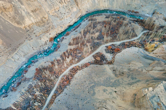 Aerial view of a vibrant turquoise river snaking through the rugged, earthy terrain of Chapursan Valley, a pathway hugging its curves, Hunza Nagar, Gilgit, Pakistan.