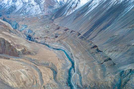 Aerial view of a winding river cutting through the rugged, mountainous terrain of Chapursan Valley, etched with pathways, against the backdrop of snow-capped peaks, Hunza Nagar, Gilgit, Pakistan.