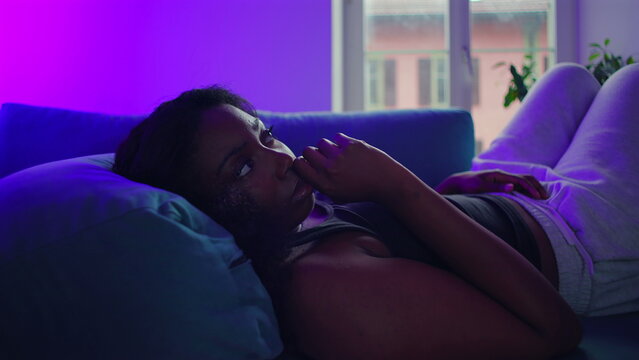African American woman lying on couch looking toward camera with thoughtful expression, hand near mouth, emotional introspection and quiet tension within intimate home environment