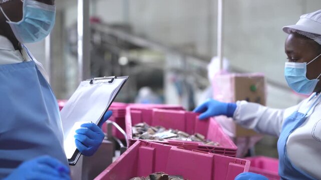 food safety researchers in lab coats discussing quality audit findings in seafood processing facility. A food safety inspector holding a clipboard while overseeing factory workers processing seafood