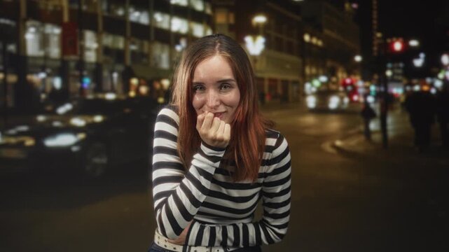 Redhead woman biting her knuckle with hand to mouth gesture on a city street at night; shyness introspection.