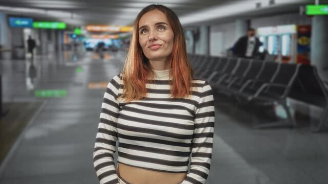 Redhead woman with hands in pockets wearing a striped cropped shirt standing in an airport concourse by empty seats; calm travel reflection.