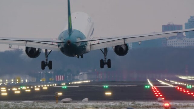 Rear view of a passenger airplane landing on an illuminated runway. The aircraft touches down, deploys spoilers, and slows down. A city skyline is visible in the background on a hazy winter day