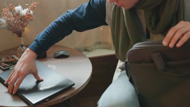 Caucasian man packs laptop into bag at wooden table, wearing green scarf and glasses, candle and dried flowers nearby, careful hands closing zipper on sleeve, relaxed morning remote work routine,