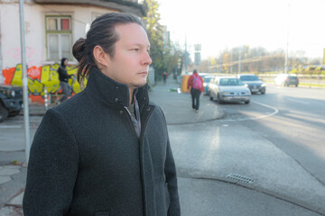 Fototapeta premium Man near busy city street with passing cars, bright sunlight, colorful graffiti wall behind, urban sidewalk, calm stance, dark coat, soft highlights, morning city movement, everyday lifestyle moment.