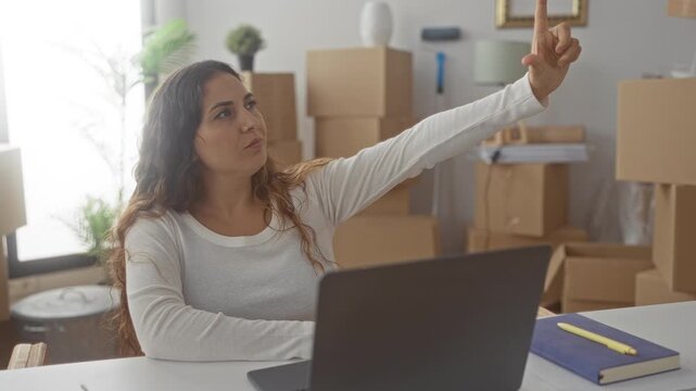 Woman at laptop pointing finger upward and hand to chin beside stacked moving boxes in apartment; anticipation.